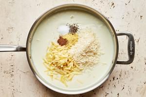 mustard powder, salt, pepper, nutmeg, Parmesan, and Gruyère being added to a roux in a skillet