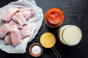 raw chicken thighs on parchment paper next to polenta, marinara, salt and chicken stock