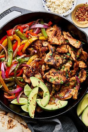 chicken fajitas with peppers and onions in a skillet topped with avocado surrounded by tortillas & queso fresco for topping