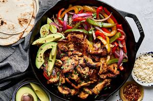 chicken fajitas with peppers and onions in a skillet topped with avocado surrounded by tortillas & queso fresco for topping