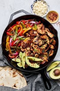 chicken fajitas with peppers and onions in a skillet topped with avocado surrounded by tortillas & queso fresca for topping