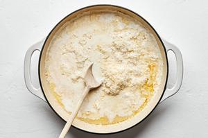 parmesan, cream, butter, garlic, garlic powder, nutmeg, salt, and pepper being stirred in a skillet with a wooden spoon