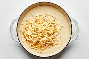 cooked fettuccine noodles being added to a pot of homemade alfredo sauce made with parmesan, cream, butter and spices