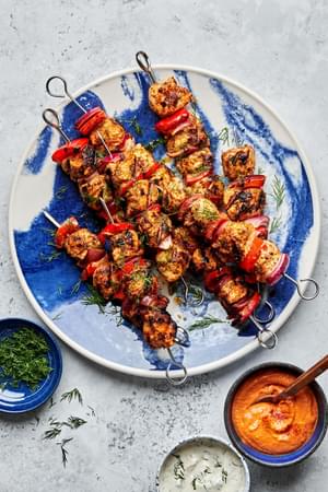 chicken kebabs with bell pepper and red onion on a serving plate served with bowls of dill sauce and romesco sauce