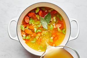 chicken broth being poured into a pot with cooked carrots, onion and celery