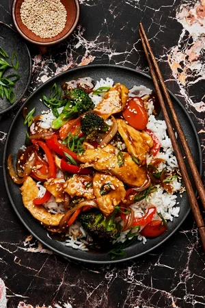 chicken stir fry with broccoli, carrots, onion & peppers on top of white rice sprinkled with green onions and sesame seeds