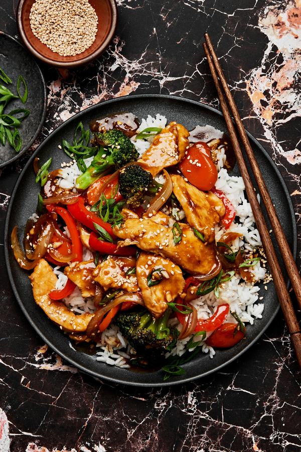 chicken stir fry with broccoli, carrots, onion & peppers on top of white rice sprinkled with green onions and sesame seeds