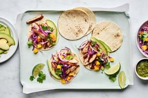 chicken street tacos with mango slaw being assembled on top of corn tortillas, garnished with avocado and lime wedges