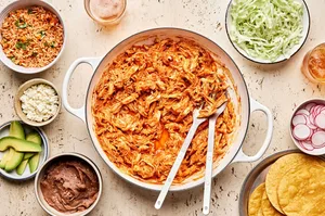 A pot filled with smoky chicken tinga surrounded by fixing to make tacos: corn tortillas, avocado, and shredded cabbage