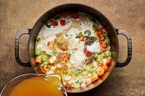 broth and cream being poured into a pot with onion, carrots, celery, garlic and butter