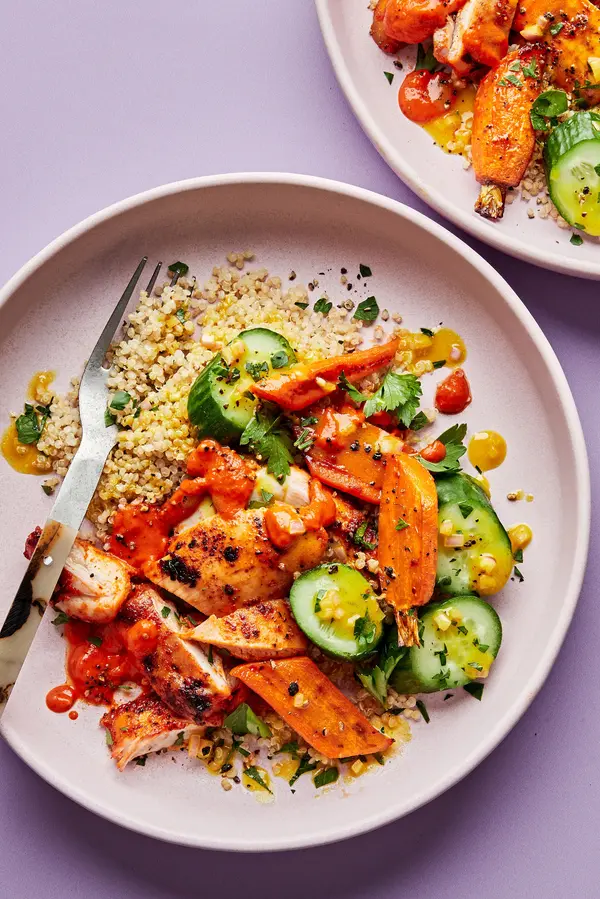 a bowl of quinoa topped with chicken with red pepper sauce, roasted carrots, cucumbers and drizzled with turmeric vinaigrette