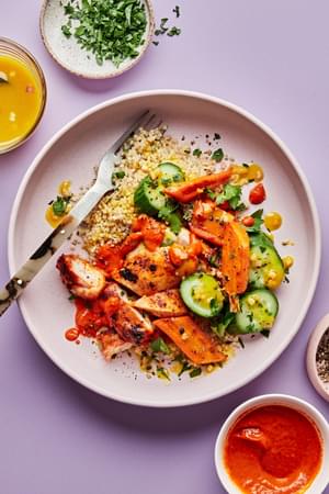 a bowl of quinoa topped with chicken with red pepper sauce, roasted carrots, cucumbers and drizzled with turmeric vinaigrette