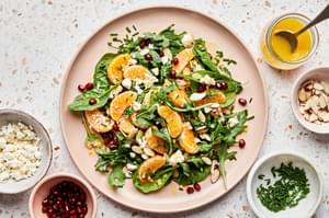 Christmas Salad being assembled with spinach, arugula, feta, clementines, pomegranates, almonds and chives