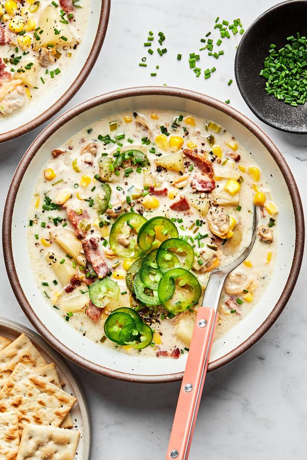 A bowl of homemade clam chowder with a spoon topped with chives next to a plate of saltine crackers