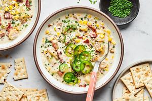 a bowl of homemade clam chowder with a spoon topped with sliced jalapeños and chives next to a plate of saltine crackers