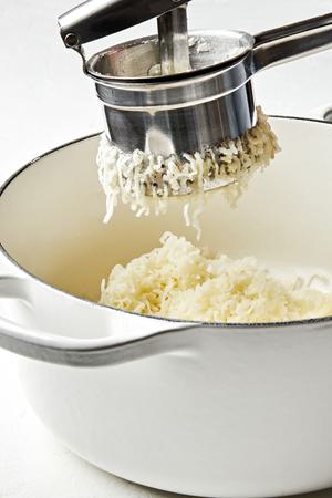 boiled potatoes being riced through a potato ricer into a large pot of warmed butter and whole milk