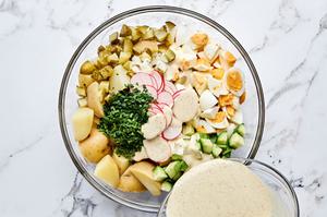 dressing being poured over potatoes, chives, parsley, cucumbers, pickles, eggs and radishes to make potato salad