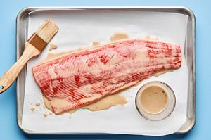 raw salmon filet being brushed with a coconut milk dressing on a parchment lined baking sheet