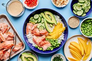 coconut rice salmon bowl surrounded by toppings: mango, edamame, cucumber, avocado, sesame seeds and coconut milk dressing