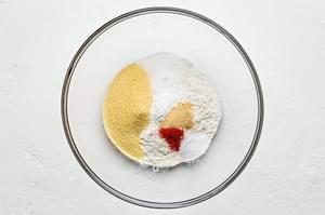 flour, cornmeal, sugar, baking powder, salt, cayenne, and garlic powder being added to a bowl