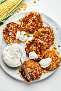 a plate of homemade corn fritters sprinkled with flaky salt served with a bowl of sour cream