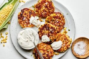a plate of homemade corn fritters sprinkled with flaky salt served with a bowl of sour cream