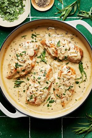 Creamy tarragon chicken in white skillet surrounded by small bowls of black pepper and fresh tarragon leaves