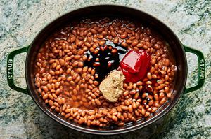 Baked beans in a pot with bacon, onion, maple syrup, spicy mustard, beef sausage, molasses, ketchup and cider vinegar.