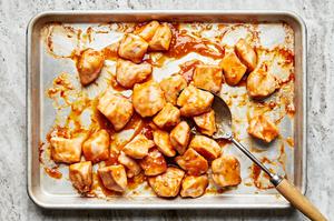 baked chicken pieces being tossed in homemade orange sauce on a baking sheet