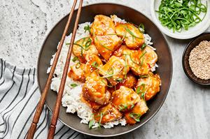a bowl of orange chicken served on top of white rice and topped with sesame seeds and green onions