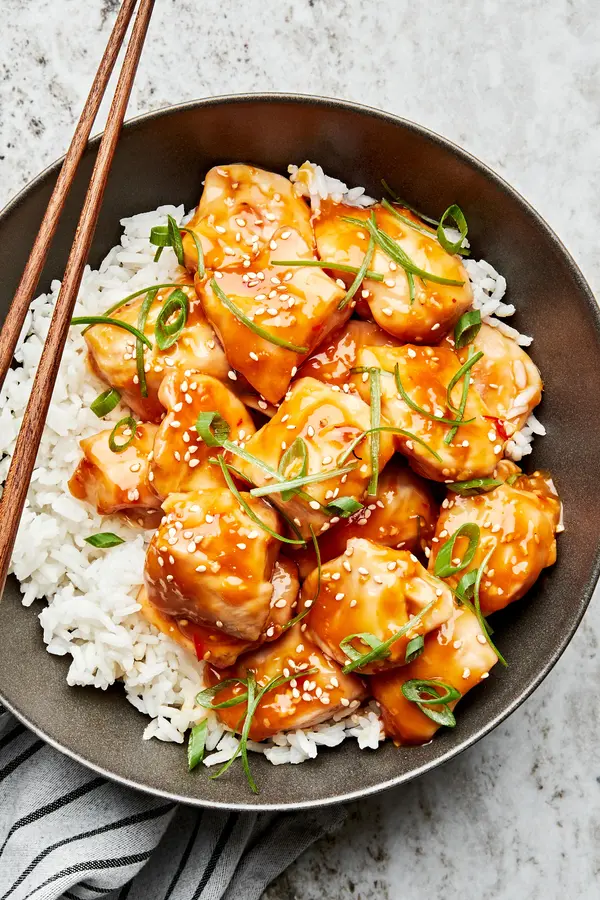 a bowl of orange chicken served on top of white rice and topped with sesame seeds and green onions