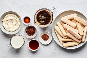 Tiramisu ingredients laid out in bowls: mascarpone, ladyfingers, espresso, whipped cream, cocoa powder, and vanilla.