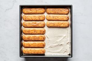 tiramisu being layered in a pan with layers of coffee soaked lady fingers and whipped spiced mascarpone