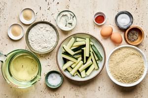 Ingredients for crispy fried zucchini: zucchini, flour, breadcrumbs, eggs, garlic powder & seasonings arranged on a counter