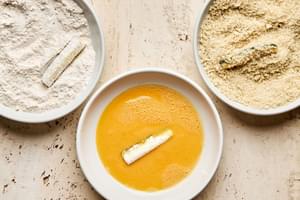 zucchini spears being dipped into bowls of flour & spices, beaten eggs, & panko to make fried zucchini