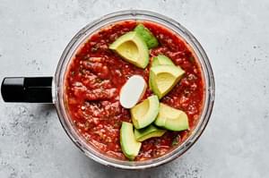 avocado being added to a food processor with charred vegetables, crushed tomatoes, cilantro, salt and white onion