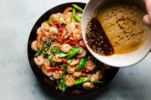 hands pouring garlic sauce over shrimp stir fry in pan.