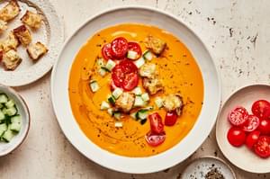 a bowl of gazpacho soup topped with a drizzle of olive oil, cherry tomatoes, cucumbers, croutons, and pepper
