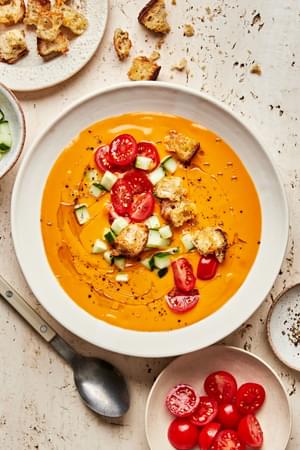 a bowl of gazpacho soup topped with a drizzle of olive oil, cherry tomatoes, cucumbers, croutons, and pepper