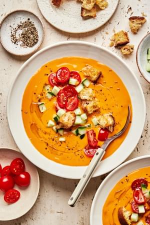 a bowl of gazpacho soup topped with a drizzle of olive oil, cherry tomatoes, cucumbers, croutons, and pepper