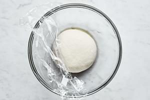gluten free pizza dough ball in a glass bowl being covered with saran wrap