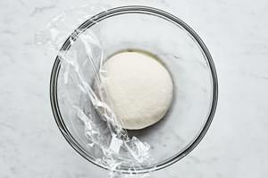gluten free pizza dough ball in a glass bowl being covered with saran wrap
