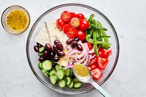 greek dressing being drizzled over tomatoes, cucumbers, bell peppers, kalamata olives and feta in a large bowl