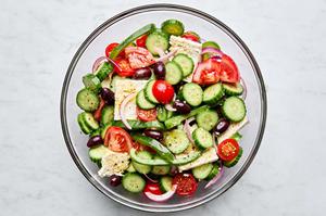greek salad being tossed in a glass bowl made with tomatoes, cucumbers, bell peppers, kalamata olives and feta