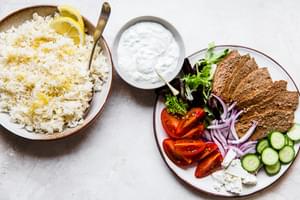 lemon rice, tzatziki sauce in a bowl and a plater with gyro meat, cucumbers, tomatoes, red onions, feta and lettuce.