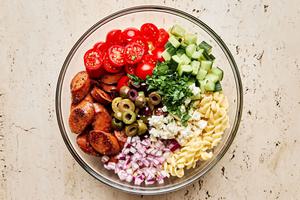 Italian sausage, pasta, greek olives, parsley, red onion, feta, tomatoes, and cucumbers being combined in a bowl