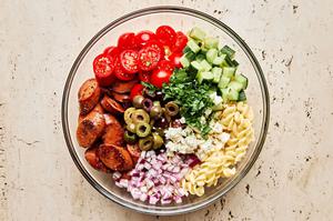 Italian sausage, pasta, greek olives, parsley, red onion, feta, tomatoes, and cucumbers being combined in a bowl