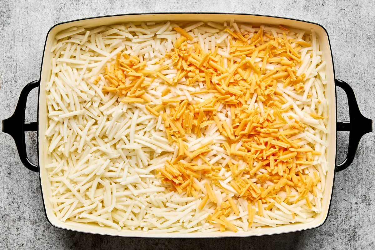Hashbrown casserole being assembled in a baking dish, with shredded potatoes, creamy sauce, and shredded cheese being layered