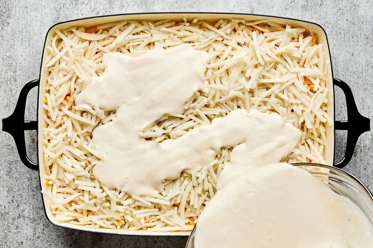 Hashbrown casserole being assembled in a baking dish, with shredded potatoes, creamy sauce, and shredded cheese being layered
