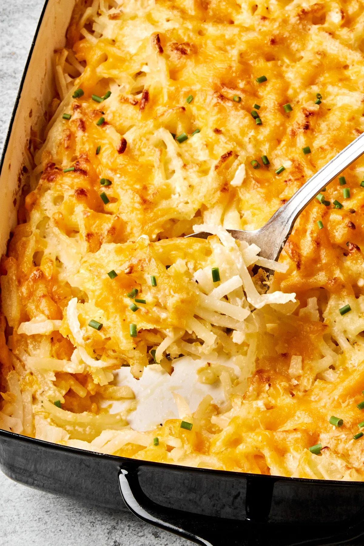 A golden-brown hashbrown casserole in a baking dish, with a creamy potato filling topped with melted cheese and chives